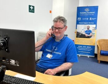 A member of the Visibility Scotland admin team sits behind a desk speaking on the phone and smiling. He is wearing a blue Visibility Scotland top.