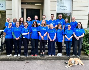 Visibility Scotland staff stand outside the main office building smiling and wearing blue Visibility Scotland tops. Gina the guide dog lays in front.