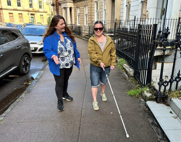 A person using a guide cane walks down a street lined with parked cars while a Vision Rehabilitation Specialist looks on.