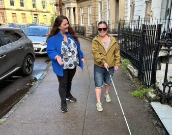 A person using a guide cane walks down a street lined with parked cars while a Vision Rehabilitation Specialist looks on.