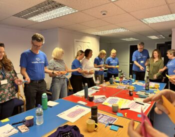 The Visibility Scotland staff team participating in a training activity around a large table. The staff are passing each other coloured post-it notes as part of a teambuilding exercise.