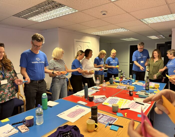 The Visibility Scotland staff team participating in a training activity around a large table. The staff are passing each other coloured post-it notes as part of a teambuilding exercise.