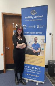 Alison smiles next to a Visibility Scotland banner. She is wearing red-tinted glasses and a black dress