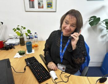 A person smiles while speaking on the phone. They are seated at a desk.