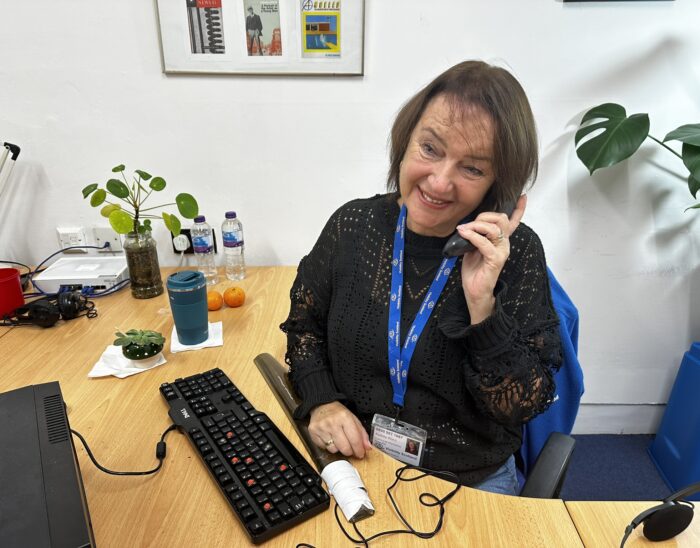 A person smiles while speaking on the phone. They are seated at a desk.