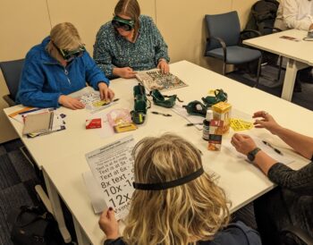 Four people sit around a table attempting to read with sim specs on
