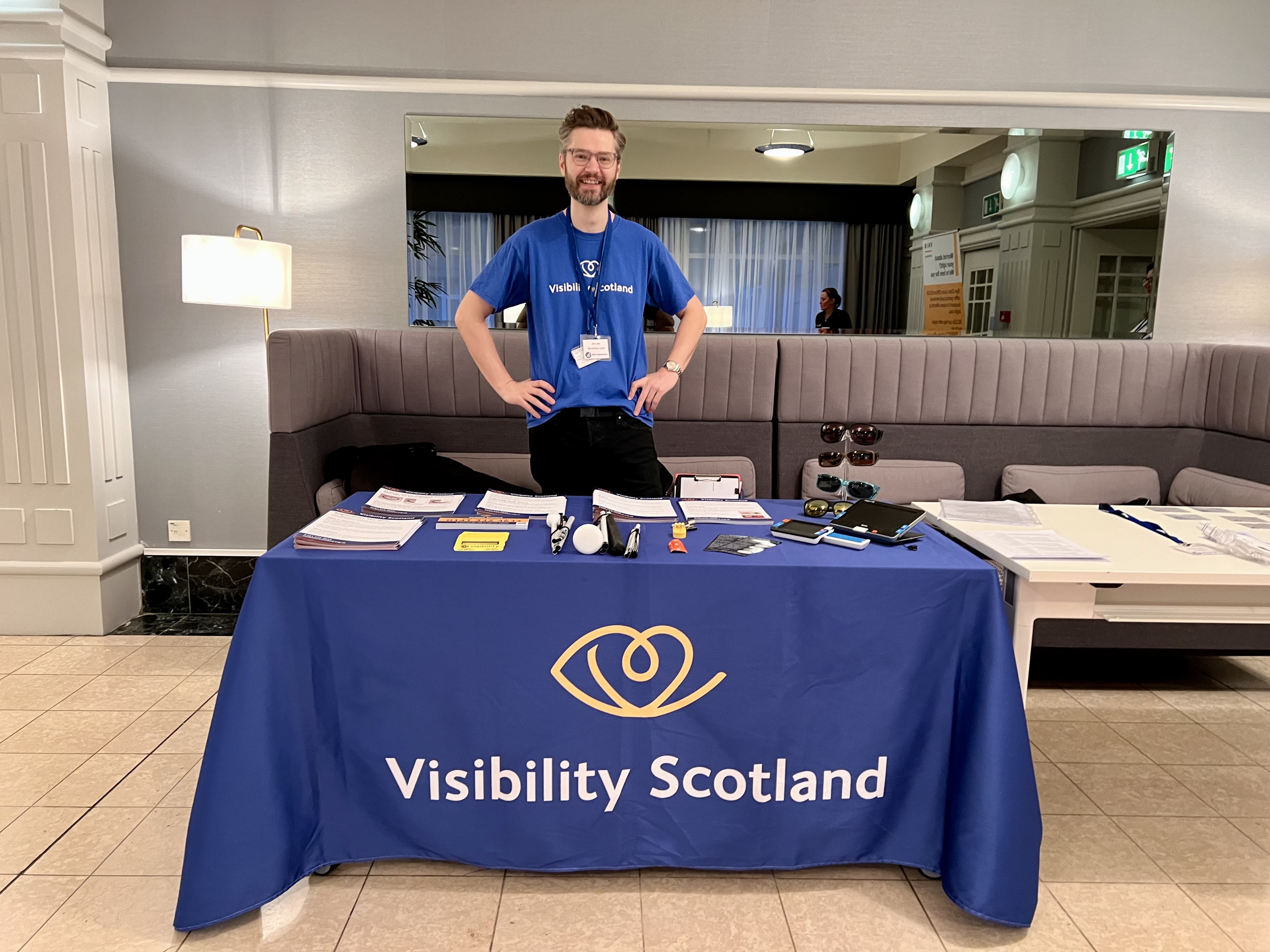 A person stands at a table with educational materials covered in a Visibility Scotland tablecloth