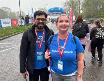 Two people stand together smiling at the Kiltwalk finish line. They are wearing Visibility Scotland tops.
