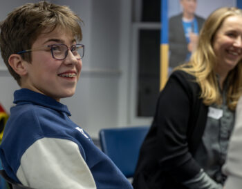 A young person smiles in focus in the foreground, in the background an adult smiles.