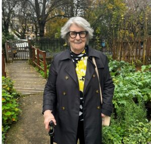 A person in a black coat and glasses stands in a garden smiling. 