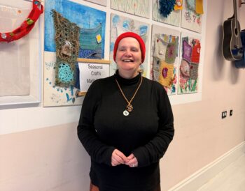 A person stands in front of a wall full of artwork smiling.
