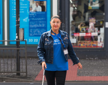 A person using a guide cane walks confidently across a road.