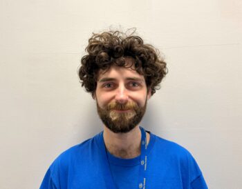 A person with brown curly hair and a beard wearing a blue Visibility Scotland top smiles at the camera