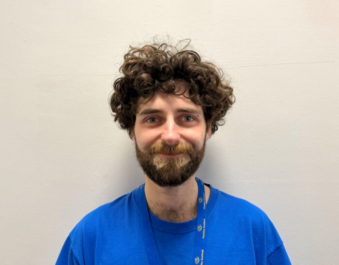 A person with brown curly hair and a beard wearing a blue Visibility Scotland top smiles at the camera