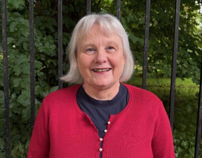 Anne stands outside smiling at the camera. She is wearing a red jumper.