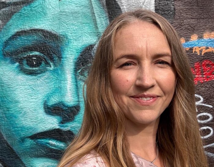 Laura stands smiling in front of an iconic Glasgow mural of a woman painted in shades of blue