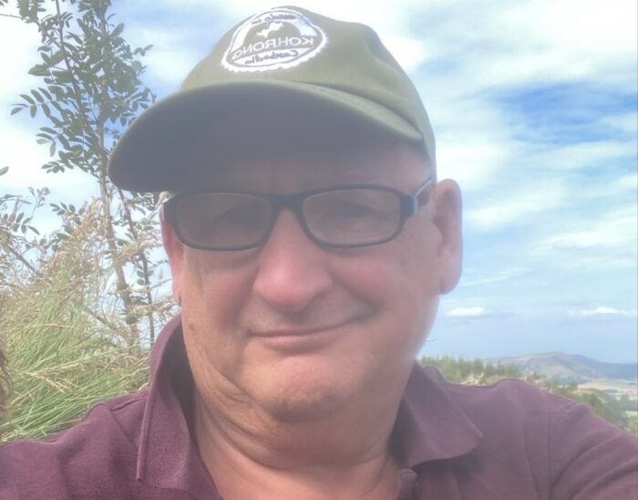 A person wearing a baseball cap, glasses, and a burgundy top smiles at the camera. They are outdoors and the sky is partly cloudy.
